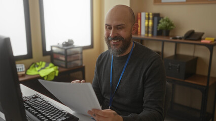 Fototapeta premium Man with beard reviewing documents at desk in modern office featuring computer monitor and shelves in background with books and decor creating a professional workspace atmosphere.