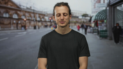 Young hispanic man with long hair standing with eyes closed and a slight smile on a busy city street outdoors, wearing a black t shirt; calm reflection.