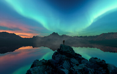 Fototapeta premium Hiker On Rocky Outcrop Under Aurora Borealis Over Mountain Range With Lake Reflection