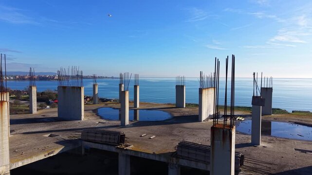Wide view of a large concrete rooftop with numerous vertical pillars. Water puddles reflect the blue sky. Sea and coast visible in the background. Aerial view.