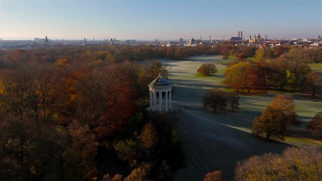 Drone footage of Englischer Garten in Munich at dawn, capturing Monopteros and colorful autumn trees. In distance are the towers of Frauenkirche, Theatinerkirche and the Bavarian State Chancellery