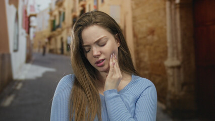 Fototapeta premium Young woman in blue sweater holding cheek with hand on old town street showing pained expression; toothache distress.