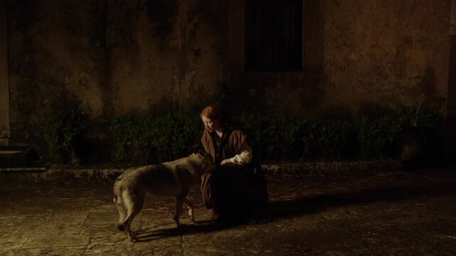 A woman in a long dress pet dogs in a quiet courtyard at night. The courtyard has old stone walls and some plants around. The moment captures a strong bond between human and dog.