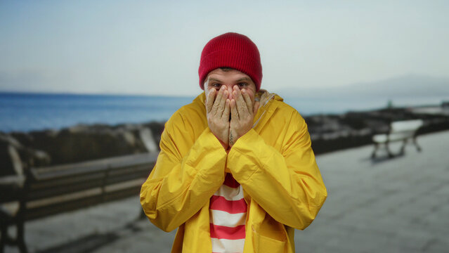 Young man with red beanie and yellow raincoat expressing joy on seaside promenade with ocean in background.