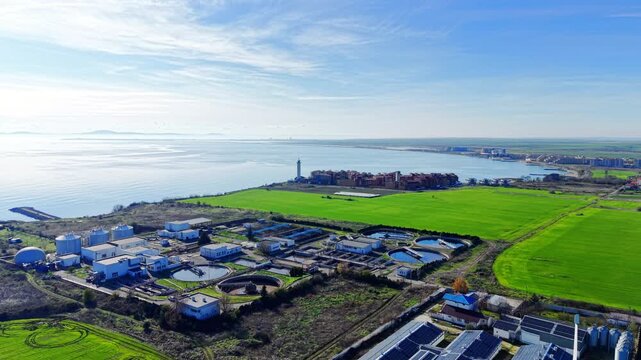 Distant aerial shot of a water treatment facility situated on a peninsula with green fields, the sea, and a lighthouse in the background.