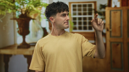 Young hispanic man in yellow shirt points finger to air in living room building while looking to the side; curiosity exploration.