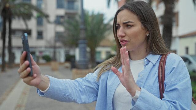 Young woman outdoors on street, using smartphone, gesturing during video call, dressed casually, showcasing urban lifestyle, engaging in digital communication with vibrant backdrop.