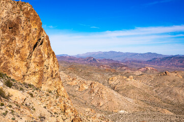 Fototapeta premium Panoramic desert landscape with rugged rocky cliffs, arid rolling hills and distant mountains under a vivid clear blue sky—sunlit, expansive vista.
