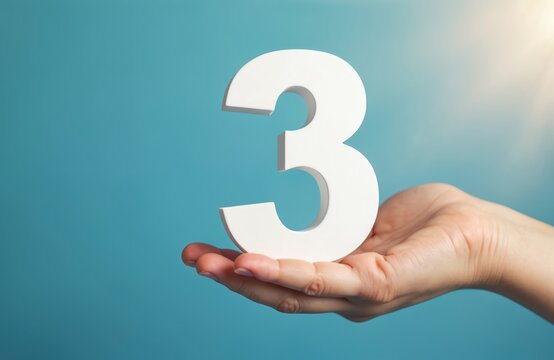 Person holds big white number three with hand. Simple clean blue background, bright light highlights the digit. Concept for third place or value.