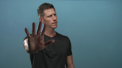 Naklejka premium Man facing camera with outstretched hand showing open palm stop gesture in a studio with blue backdrop and plain black t shirt; defiance.