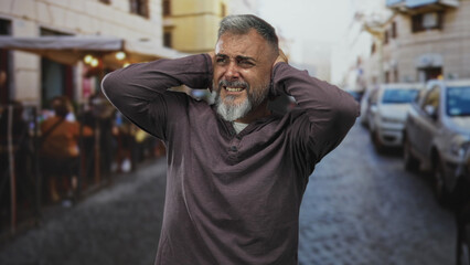 Obraz premium Man covering ears with hands and wincing on a street terrace outside a restaurant; noise discomfort.