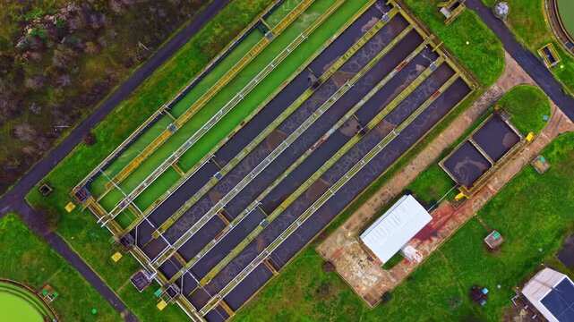 A wastewater treatment facility is shown from above, displaying several process tanks, walkways, and green patches of grass surrounding the structures in the area.