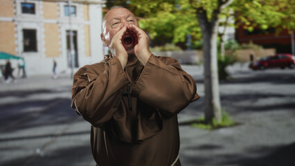 Obraz premium Senior hispanic man monk in brown habit raises both hands to his face and gestures while wearing a cross necklace on street; devotion.