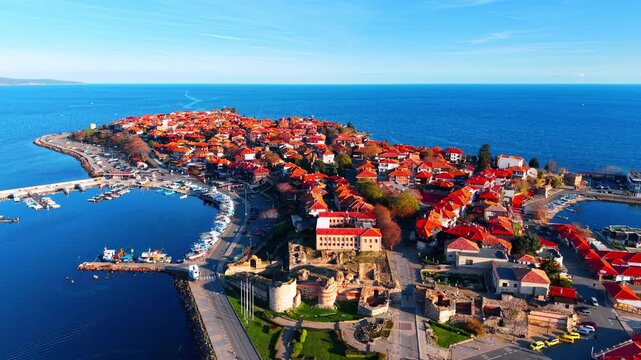 Panoramic drone shot of the ancient peninsula of Nessebar, showing traditional Bulgarian architecture, red tiled roofs, fortress ruins, and harbor.