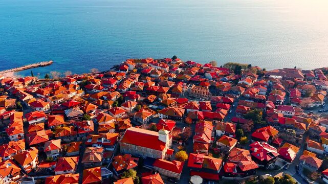 Densely packed traditional houses with orange and red tile roofs in a historic European coastal town, overlooking the calm sea surface.