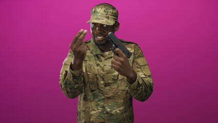 Obraz premium Man soldier in camouflage uniform holding a gun to his head while making a pinching fingers gesture against a magenta studio backdrop, smiling; tension threat.