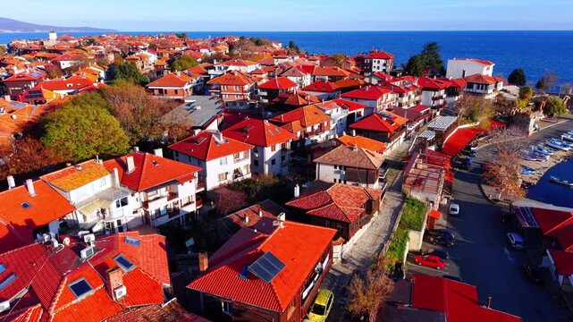 High angle drone shot of traditional houses with red clay tile roofs. Dense urban architecture of an old European peninsula town surrounded by the sea. Bright daylight.