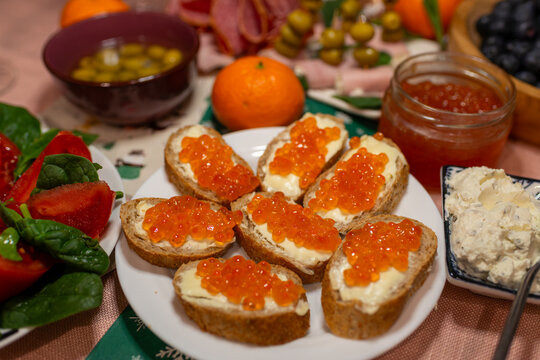 Plate of salmon roe canapes on sliced bread with butter, surrounded by olives, blueberries, tangerine, and spreads on a festive table. Gourmet party appetizer concept with bright orange caviar and hol