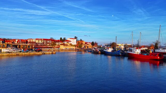 Aerial view of a harbor with boats and old town. Coastline with traditional buildings and a row of colorful fishing boats docked. Blue sky with white contrails.