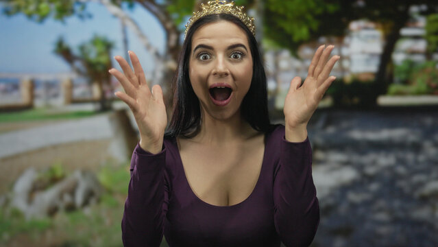 Young hispanic woman with crown expresses surprise in an outdoor park filled with trees and sunlight, enhancing her joyful demeanor and vibrant atmosphere.