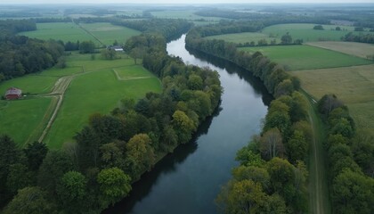 Fototapeta premium Aerial view of wide river flowing through green fields and lush forests. Rural landscape with trees, meadows, and distant hills. Peaceful natural scenery on a cloudy day.
