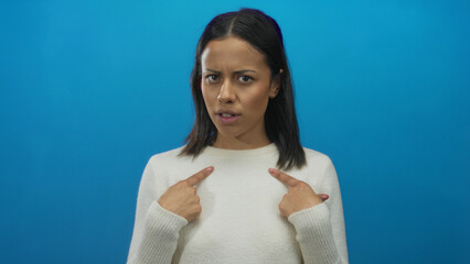 Obraz premium Woman pointing to herself with a questioning expression against a solid blue background, conveying curiosity and self-awareness, dressed in a white sweater.
