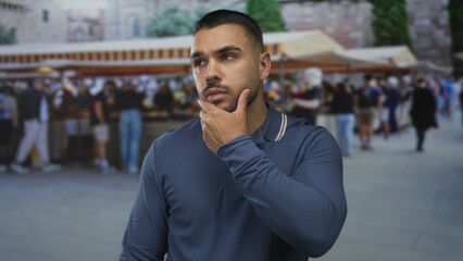 Fototapeta premium Man wearing navy shirt with subtle pensive expression touches chin in street market framed by historic building facades; thinking.