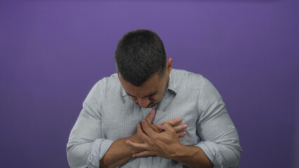 Obraz premium Young hispanic man clutching chest with pained expression in purple studio; pain distress heartattack emergency.