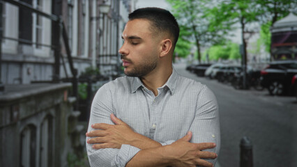 Fototapeta premium Young hispanic man in gray shirt crossing arms on city street lined with parked cars and leafy trees; rejection.
