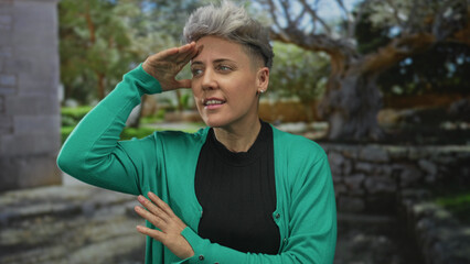 Fototapeta premium Woman with short silver hair shading her eyes with hand amid sunlit trees and stone walls in forest setting; curiosity exploration.