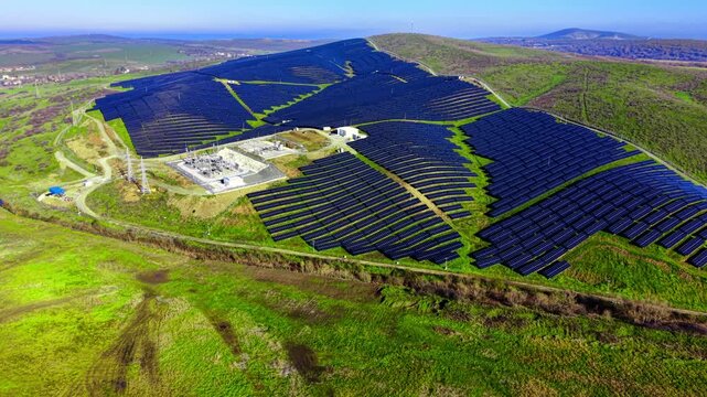 Wide aerial panorama of a massive solar power plant covering hillsides. A central electrical substation with transformers and power lines. Green fields and a clear horizon surround the facility.