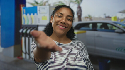 Smiling woman in striped shirt extends palm toward camera at petrol station forecourt by fuel pump and parked car  warm welcome. © Krakenimages.com