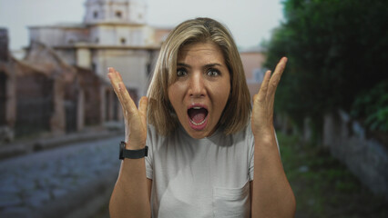 Woman with mouth agape raising both hands and screaming on cobblestone street in front of old...