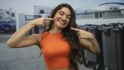 Fototapeta premium Woman in orange crop top pointing finger to bright teeth and wide smile on street next to recycling container and trash bins; happiness.