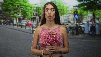 Fototapeta premium Young hispanic woman holding pink bouquet with hands on street puckers lips in fish face; playful curiosity.
