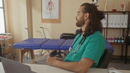 Obraz premium Man doctor with stethoscope typing on laptop at desk in clinic building, seated with hands on keyboard and focused expression; calm concentration professionalism.