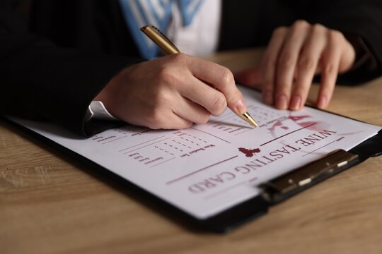 Professional sommelier filling out wine tasting card at wooden table, closeup