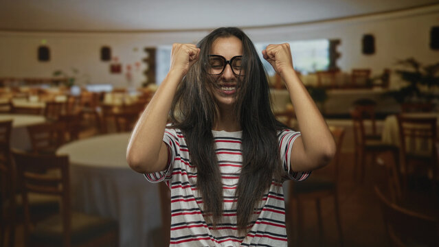 Woman wearing glasses and striped tshirt holding hair with hands and cheering in building restaurant dining room full of tables and chairs; joy celebration.