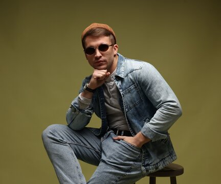 Fashion model. Handsome man in hat, sunglasses and jacket posing on stool against dark green gradient background