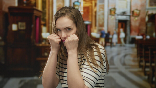 Woman holding fists raised near face inside a historic church with marble floors and wooden pews; determination.