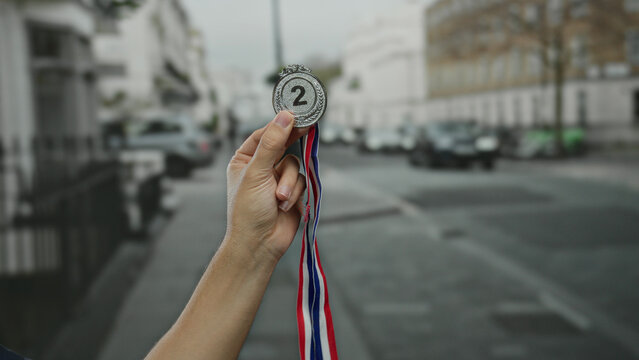 Caucasian man hand holding silver medal with ribbons on a city street, surrounded by urban outdoor scenery and buildings, symbolizing second place achievement.