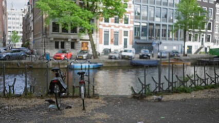 Blurred view of amsterdam canal with trees, bicycles, and parked cars under clear sky, capturing the essence of the netherlands in an urban setting.