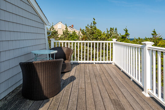 Coastal home deck with outdoor seating, wooden floor and white railing overlooking greenery on sunny day