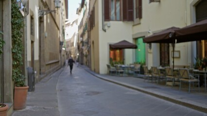 Fototapeta premium Narrow outdoor alley blurred bokeh with sidewalk cafe, tables and umbrellas, cobbled pavement and nopeople depth; background backdrop copyspace calm.