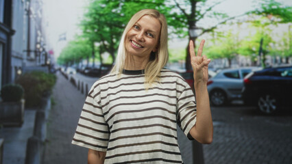 Obraz premium Blonde middle aged smiling woman holding up two fingers with right hand in peace sign on street wearing striped tshirt; cheerful.