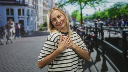 Fototapeta premium Woman smiling with outstretched hands and palms forward on a street beside a bicycle lined canal bridge, wearing a striped shirt and casual pose; warm welcome joy.