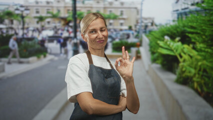 Fototapeta premium Woman in apron and white t shirt makes ok sign with right hand, arm crossed, smiling on a busy city street lined with planters and pedestrians; confidence.