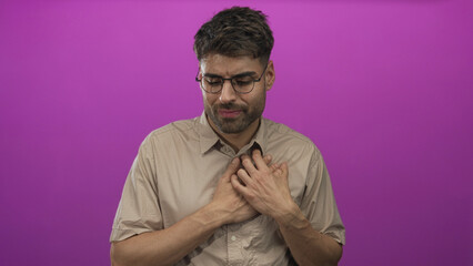 Obraz premium Young hispanic man wearing glasses and a beige shirt clutching chest in purple studio; heartattack pain.