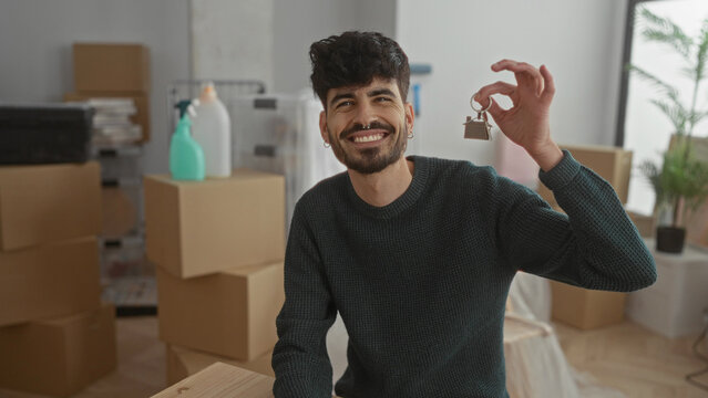 Young man smiling indoors holding keys in a new home surrounded by moving boxes and plants, representing a fresh start in an apartment setting.