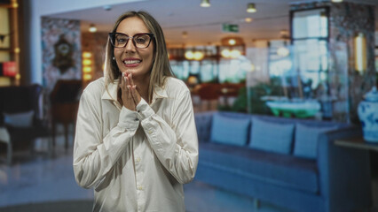 Fototapeta premium Woman hispanic young blonde clasping hands near face in hotel building lobby, smiling with glasses and white shirt and pearl earring; hope.